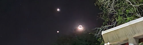 Un cielo nocturno que presenta una luna llena brillante ubicada en el centro, flanqueada por dos cuerpos celestes menos intensos y enmarcada por la silueta oscura de hojas de árboles a la izquierda y parte de un edificio a la derecha.