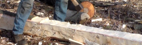 Una persona con jeans y botas de trabajo usa un hacha tradicional para partir un tronco en un día soleado, rodeada por una zona boscosa.