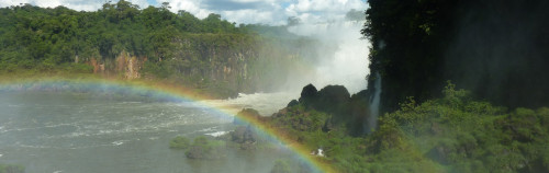 Una vista panorámica de una gran cascada en medio de una exuberante vegetación, con niebla elevándose desde la cascada y un arcoíris vibrante arqueándose sobre las aguas turbulentas de abajo.