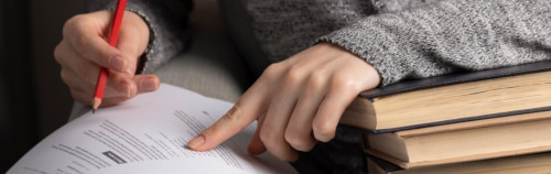Primer plano de las manos de una persona, una apuntando a un documento mientras la otra sostiene un bolígrafo rojo, con una pila de libros a su lado.