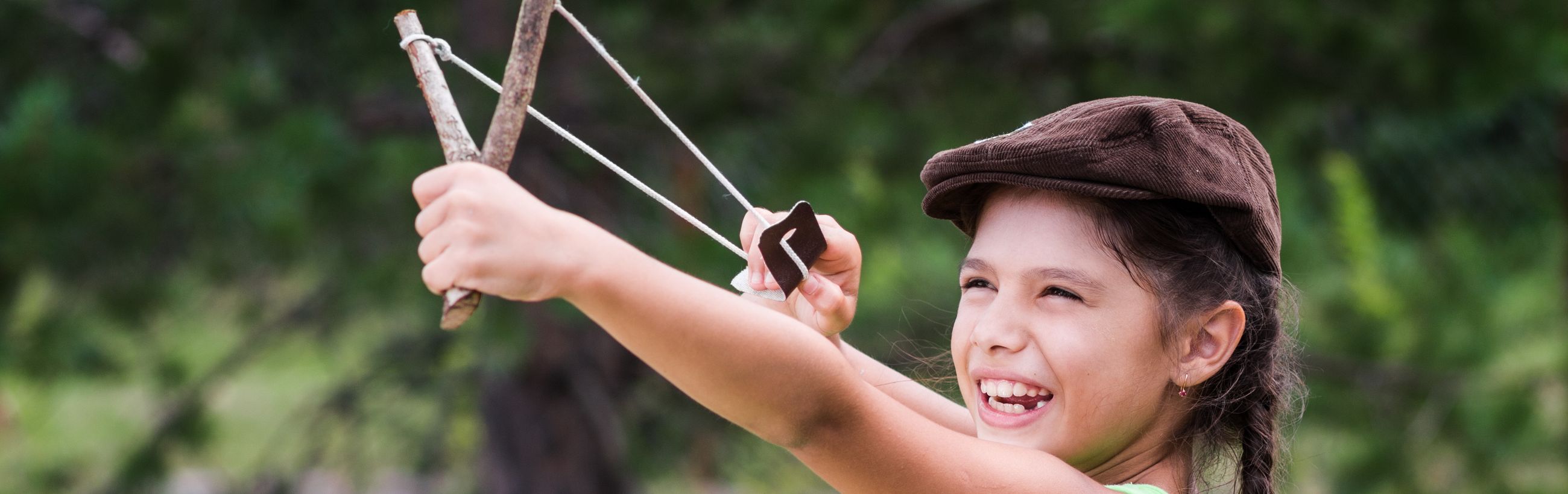 A young girl with joyful expression aiming a handmade slingshot in a lush green park, wearing a brown cap.