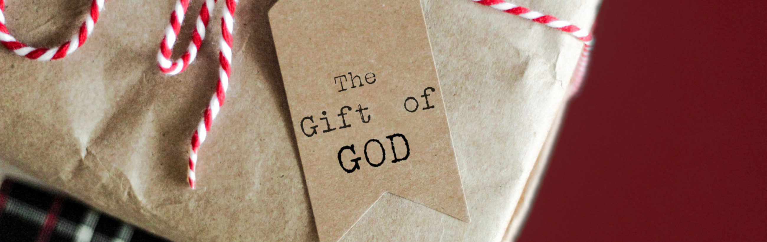 Close-up image of a craft-style gift wrapped in brown kraft paper, tied with a red and white candy-striped twine, with a tag stating 'The Gift of GOD' in black print.