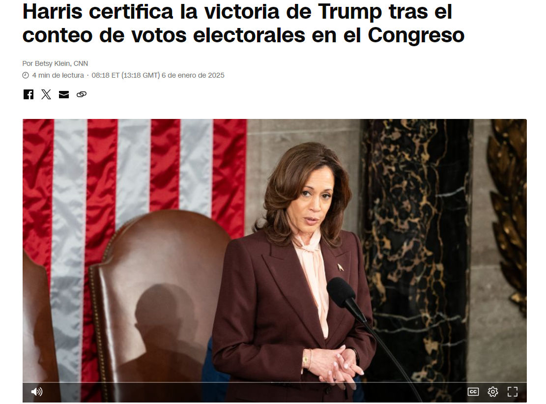 Una mujer con traje marrón habla en un micrófono frente a varias banderas estadounidenses. Está en una sala decorada con columnas de mármol oscuro y un tapiz dorado.