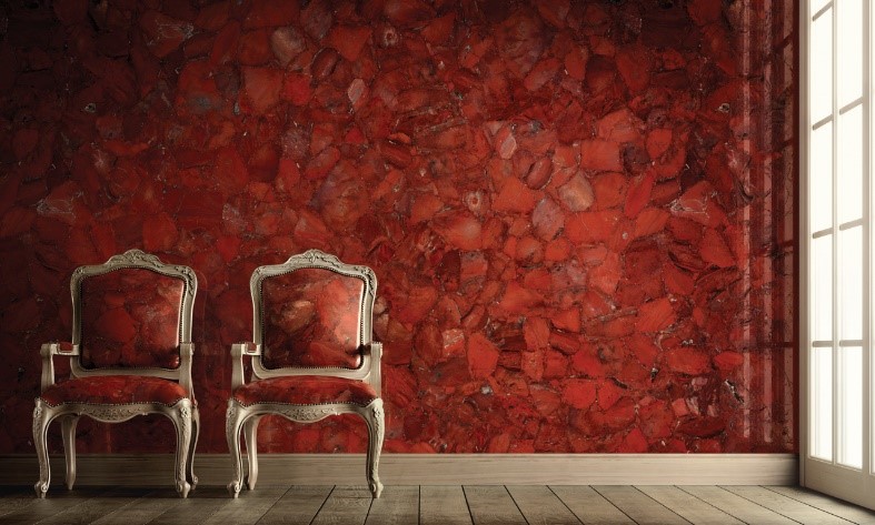 Two vintage chairs with ornate silver frames and deep red upholstery stand in front of a vibrantly textured red wall composed of various stones. Wooden flooring stretches across the room, illuminated by natural light entering through the tall window on the right.