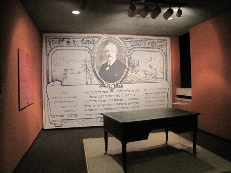 An exhibit room with dim lighting displaying a large wall mural featuring an older man's portrait encircled by ancient script and imagery that includes traditional agricultural scenes. In the foreground, a simple wooden table stands centered on a light-colored rug.