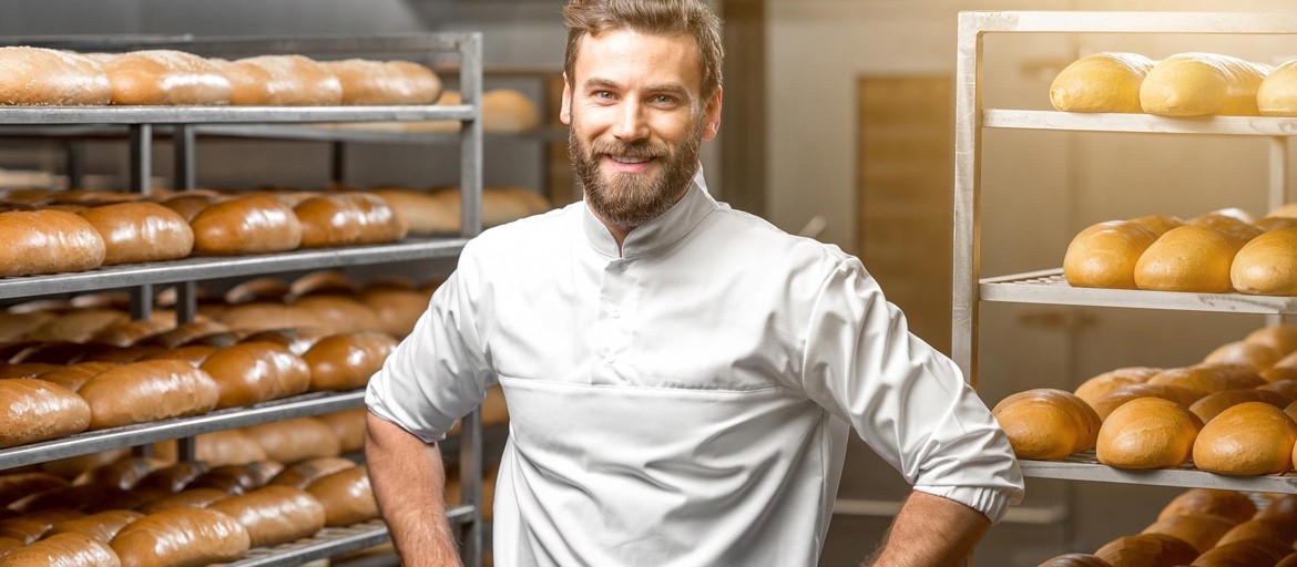 A baker in a white chef’s uniform stands confidently in a bakery, smiling with hands on his hips. Behind him, racks filled with freshly baked loaves of bread showcase various sizes and shades of golden brown crusts.