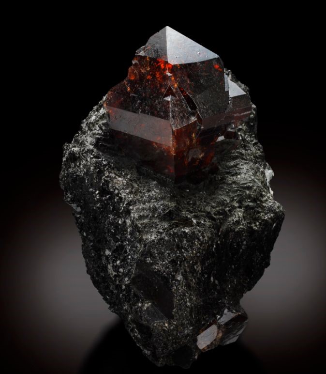 A dark, rough textured mineral hosts an angular, translucent garnet crystal with rich, red ember hues, evoking the brilliance of celestial objects in the night sky.