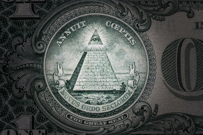 Close-up view of a detailed emblem on a dollar bill featuring an eye within a triangle above a pyramid with the Roman numerals MDCCLXXVI at the base, surrounded by woven patterns and the phrase "Annuit Coeptis Novus Ordo Seclorum."