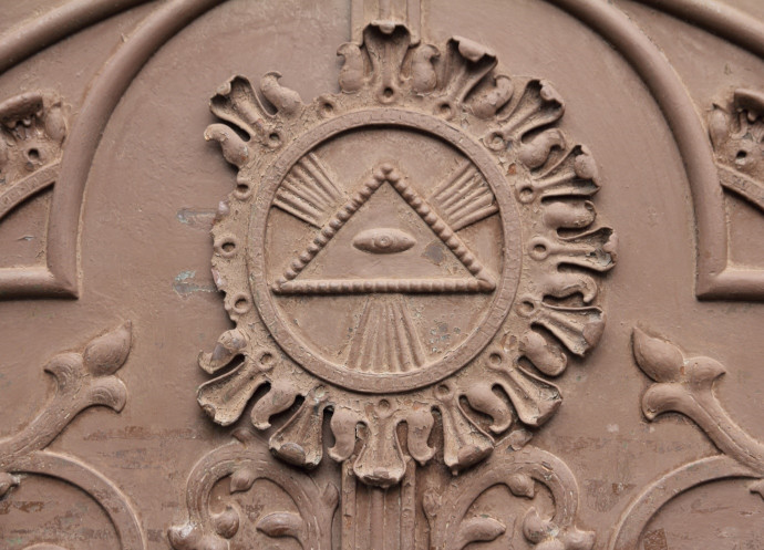 Detailed bas-relief of a triangular emblem surrounded by rays within a circular ornate frame, on a textured brown surface featuring decorative foliage and scrollwork.