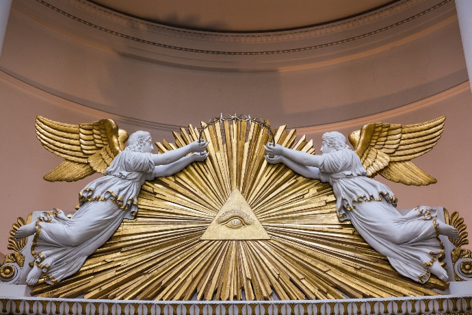 Two white sculptures of angels with golden wings, each turned towards a central golden radiating symbol featuring an eye within a triangle. This artwork is displayed against a soft, curved architectural background in subtle pastel shades.