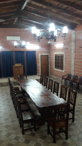 A rustic dining hall with a long wooden table and several wooden chairs under an ornate chandelier. The room features exposed wooden beams, terracotta brick walls, and tiled flooring.