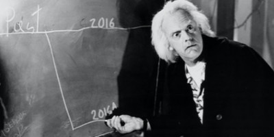 A black and white photo depicts an elderly man with wild gray hair standing in front of a chalkboard, on which a graph is drawn with the years 2016 indicated on the horizontal axis. The man appears to be in thought or mid-explanation, looking directly at the viewer with a mix of surprise and contemplation.