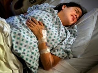A woman lying in a hospital bed appears to be in discomfort, resting with a bandaged wrist and her hands on her chest.