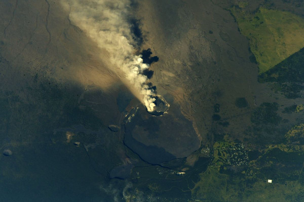 Aerial view of a smoking volcano casting a shadow upon its own ashen landscape, surrounded by verdant terrains, illustrating the dynamic creation processes of the Earth.