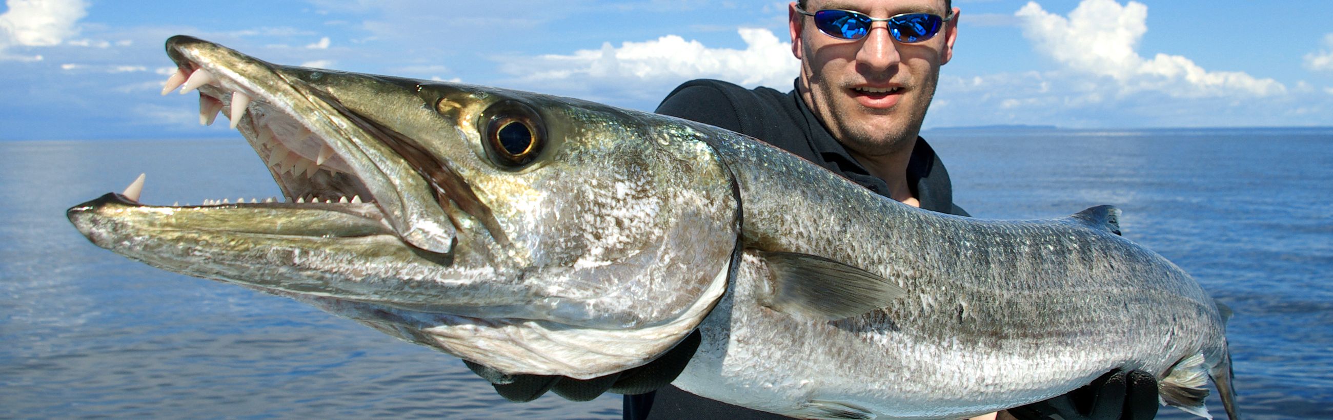 A man in sunglasses smiling broadly as he holds a large barracuda fish in both hands, displaying its sharp teeth and reflective scales. They are both in front of a serene ocean backdrop under a partly cloudy sky.