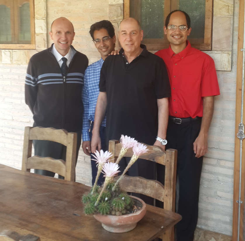 Four men of varied ages, smiling in a semi-circle behind a rustic wooden table with a clay pot containing a cactus with three pink flowers at its center. They stand in front of a stone building with timber doors, under a shaded porch.