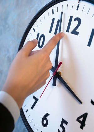 A person adjusts the minute hand on a large white analog clock with black numerals and hands, setting it to 12 o'clock.