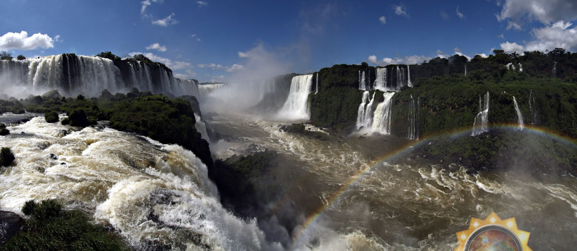 Panoramic view of a majestic waterfall system with multiple cascades plunging into a swirling river below, surrounded by lush green vegetation. A rainbow arches gracefully over the misty waters, and an illustrative overlay of a celestial chart sits at the bottom right corner reflecting the Mazzaroth.