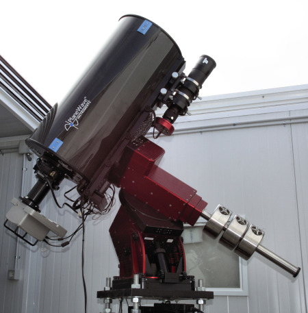 A large astronomical telescope with a shiny silver tube and a red mounting system is positioned inside an observatory with its roof partially open, pointing towards the sky for celestial observations.