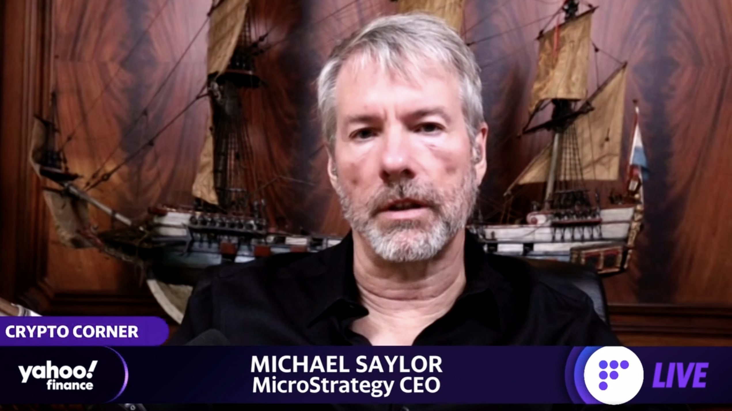 A Caucasian man in a black shirt appears on a live video with a title reading "Michael Saylor, MicroStrategy CEO." The background is a richly textured wooden panel featuring a model of a large sailing ship. The setting is part of a segment labeled "Crypto Corner" by Yahoo Finance.
