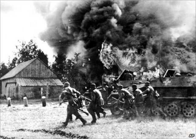 Black and white historical photo of soldiers advancing near a tank with smoke billowing from an explosion behind a building, in a rural setting.