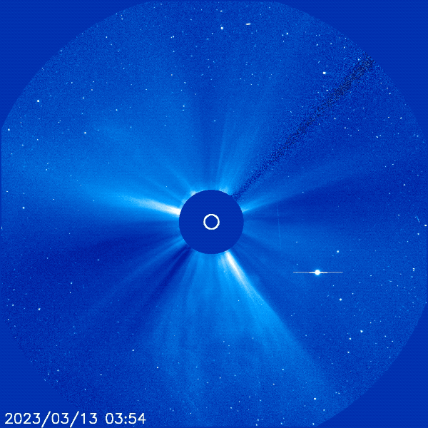 An astronomical image showing the sun blocked out by a coronagraph, creating a radial display of sunlight scattering in the solar corona. A comet is visible with a distinct tail extending towards the right, amidst numerous scattered points of light representing stars. A timestamp in the bottom left reads “2023/03/13 03:54”.