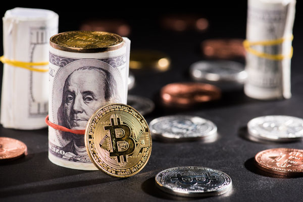 A collection of various currencies including a rolled stack of U.S. dollars bound by a rubber band, a prominently placed gold-colored Bitcoin, and assorted coins scattered on a dark surface.