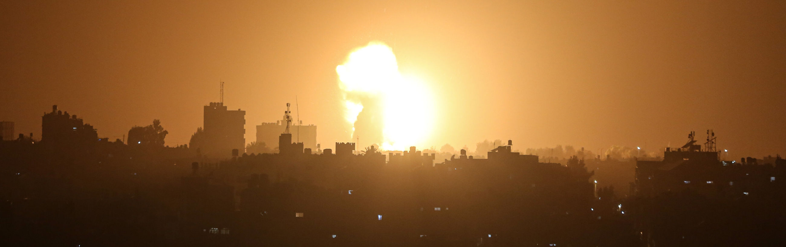 A panoramic view showing a cityscape at night under a sky glowing with a fiery orange light from a large, bright explosion on the horizon, casting a stark illumination over the silhouetted buildings.