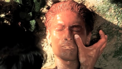 A close-up image depicting a person's face, partially covered in a golden substance, lying on the ground with another person's hand gently touching their face. The setting suggests an outdoor scene with natural light casting shadows over the facial features.