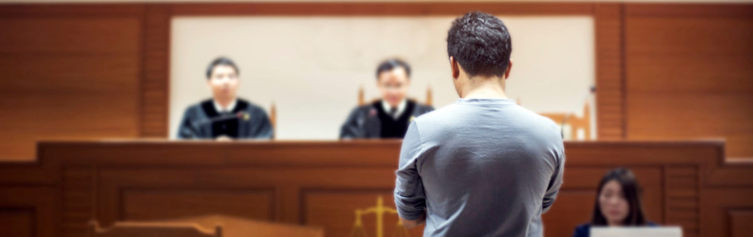 A man standing in a courtroom facing two judges who are out of focus, emphasizing the tension and focus on the individual.