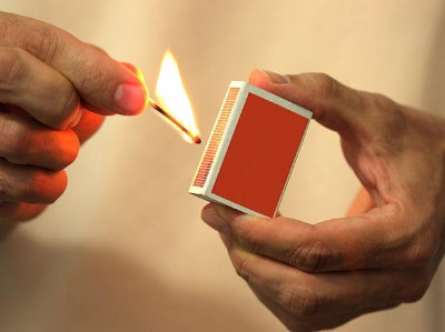 Close-up of two hands, one striking a match from a red matchbook, igniting the match head with a visible flame against a blurred background.