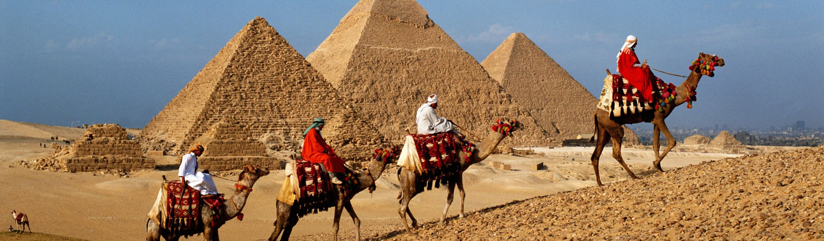 Three people riding colorfully decorated camels in front of the ancient pyramids under a clear sky.