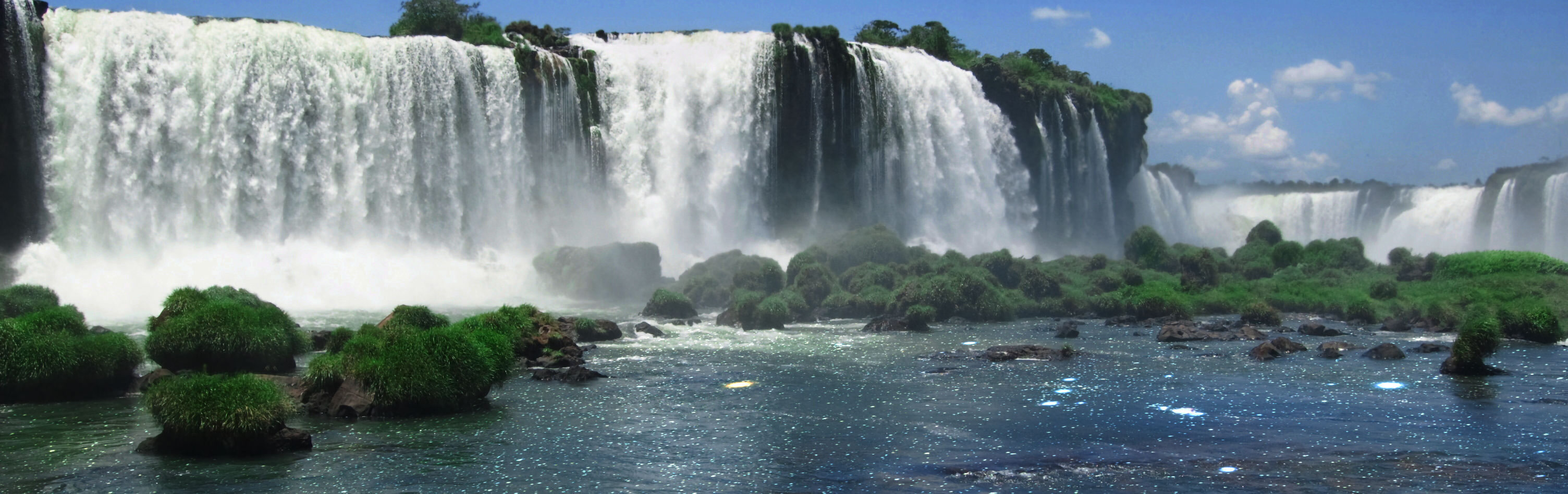 Wide panoramic view of a majestic waterfall cascading into a river, surrounded by lush greenery and scattered boulders under a clear blue sky. Mist rises from where the water violently meets the river, and the sunlight sparkles on the water's surface.