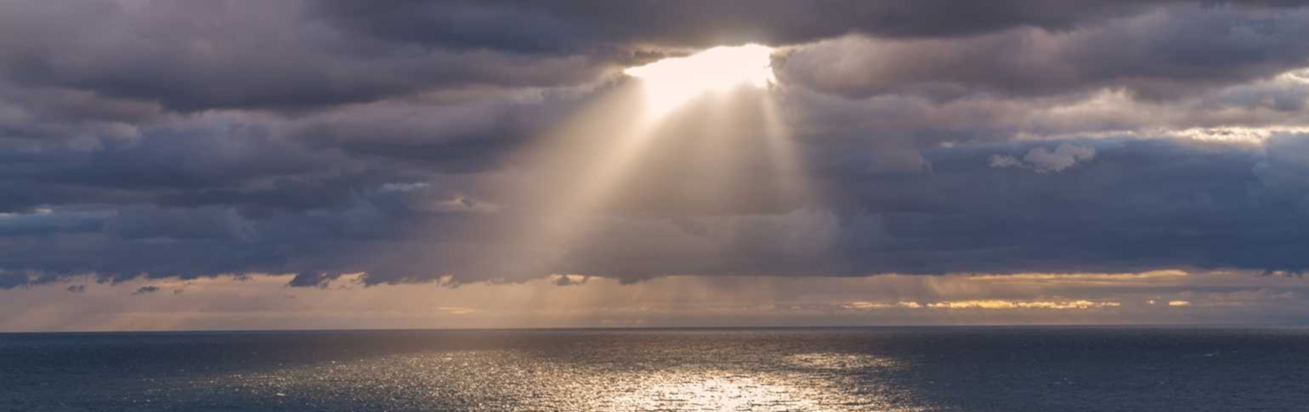 A vast ocean under a dramatic sky, where rays of sunlight break through a dense cloud cover, casting a brilliant path of light across the sea surface.
