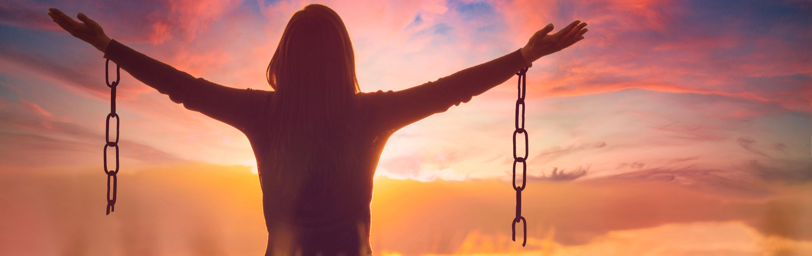 Silhouette of a person with arms raised wide and holding chains, backlit by a vibrant sunset sky with hues of orange, pink, and blue.
