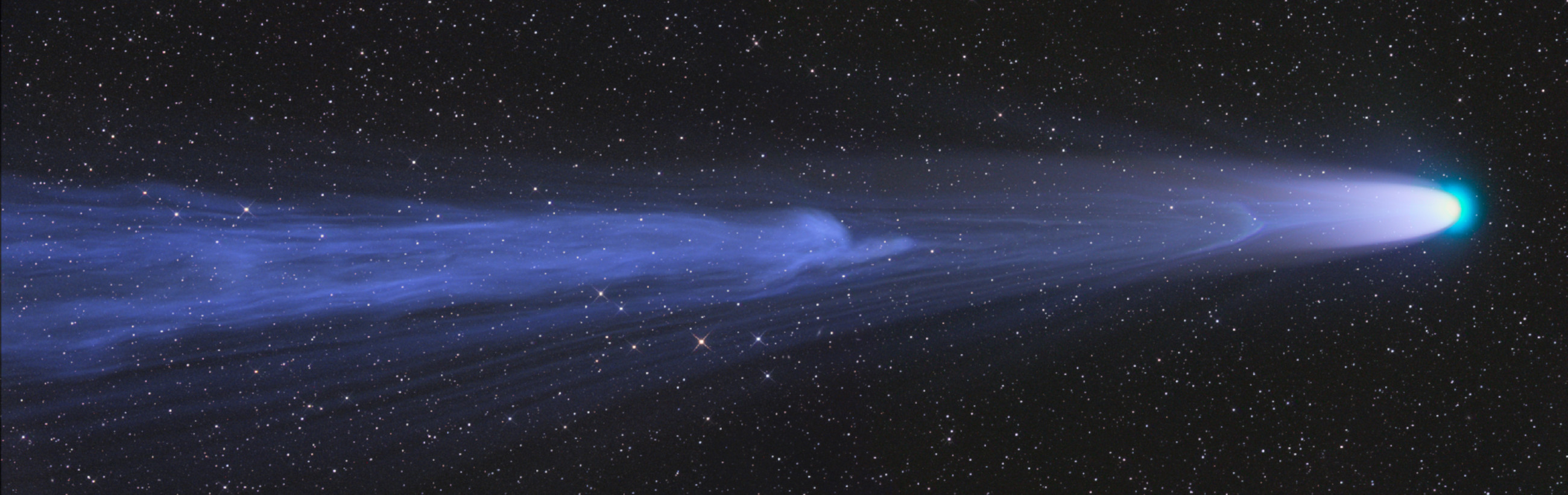 A detailed nighttime view of the sky showing a brilliant comet with a glowing blue nucleus and a long, sweeping tail made of gas and dust, against a backdrop of countless stars.