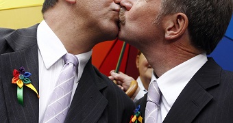 Two men in formal attire sharing a kiss. One man wears a purple tie and a multicolored ribbon pin, the other a silver tie. A rainbow flag is visible in the background, suggesting a celebration.