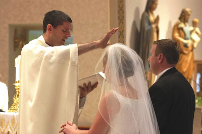 A priest in white robes blessing a bride and groom during a wedding ceremony inside a church, with religious statues visible in the background.