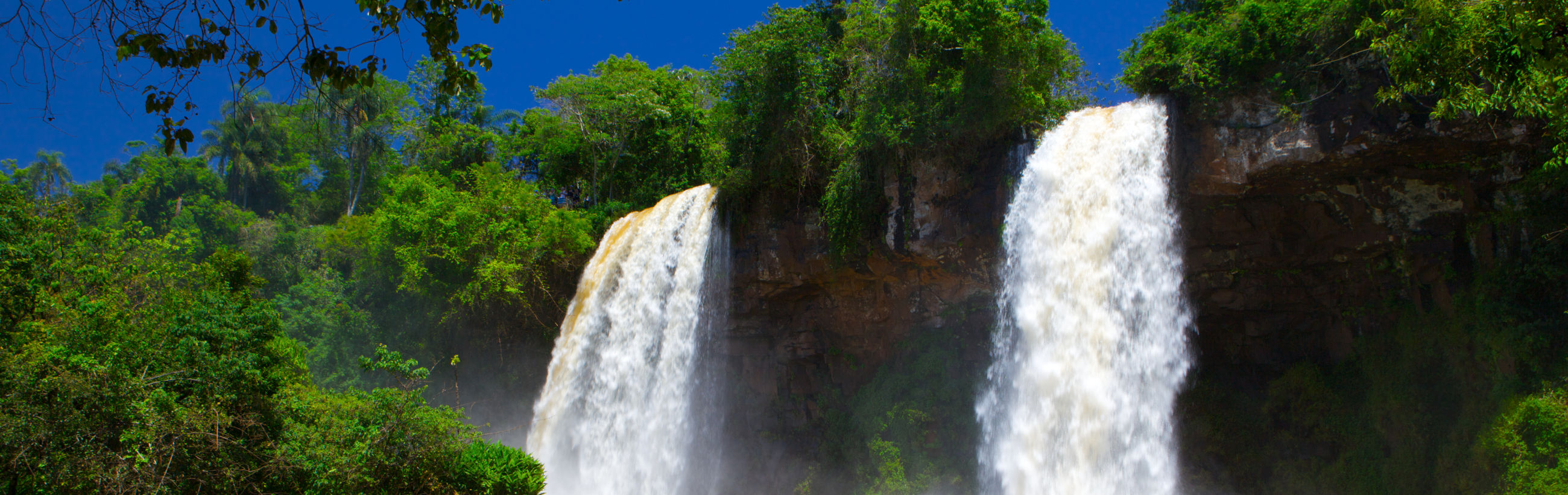 A panoramic view of a vibrant waterfall flanked by lush green foliage under a bright blue sky.