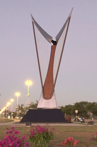 A large outdoor sculpture resembling an abstract representation of the Mazzaroth, constructed with triangular metallic frames holding stretched wires, sits in a landscaped park area with street lamps and flowers at dusk.