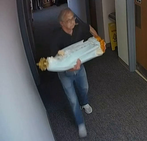 An older man walks down a narrow, dimly lit corridor carrying a large white decorative item adorned with golden ends, resembling a historical artifact.