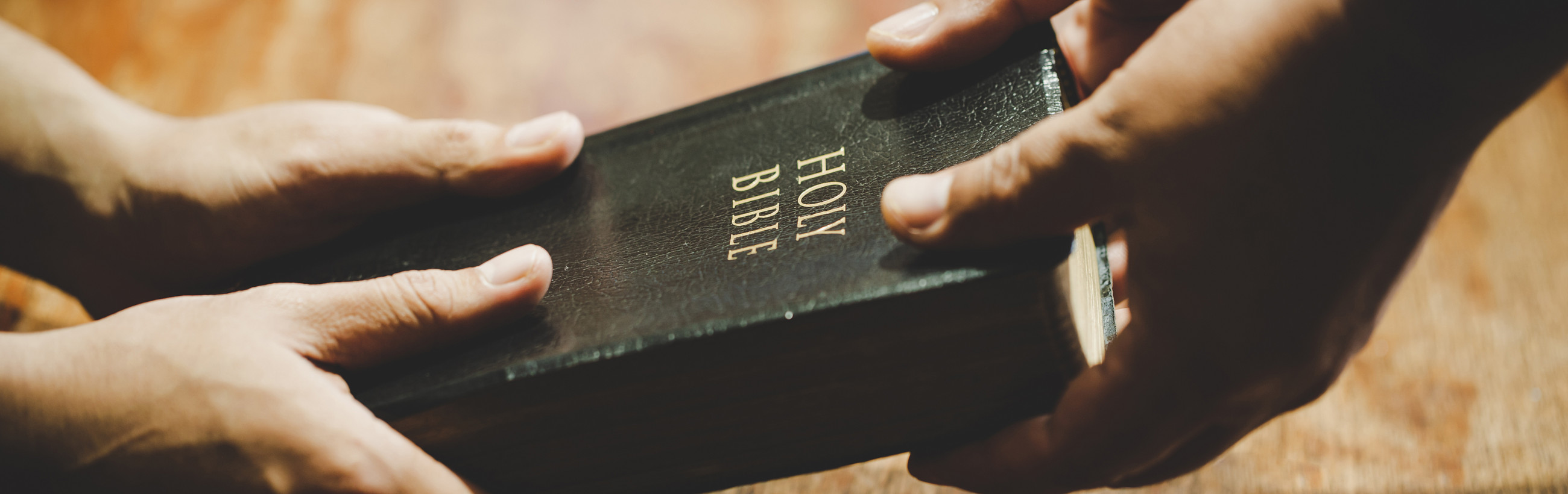 Close-up view of two hands holding a closed Holy Bible on a wooden table, emphasizing the embossed title on its cover.