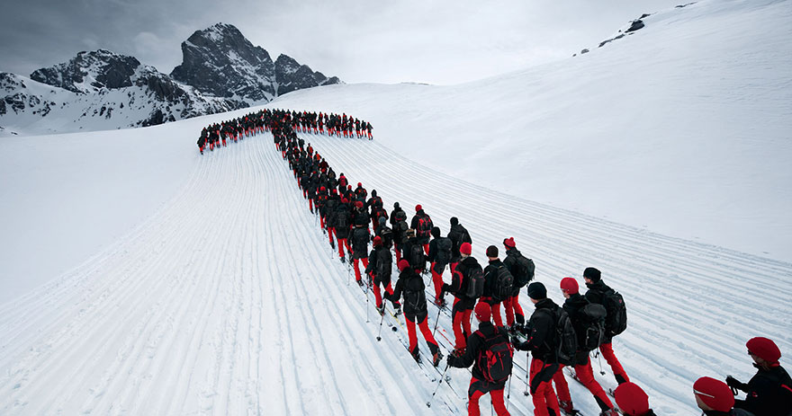 A large group of individuals clad in red jackets and black trousers are methodically trekking across a snowy mountain landscape, forming a winding line reminiscent of the Mazzaroth configuration in a clear, daylight setting. The snowy expanse and rugged mountain peak in the background enhance the dramatic scene.