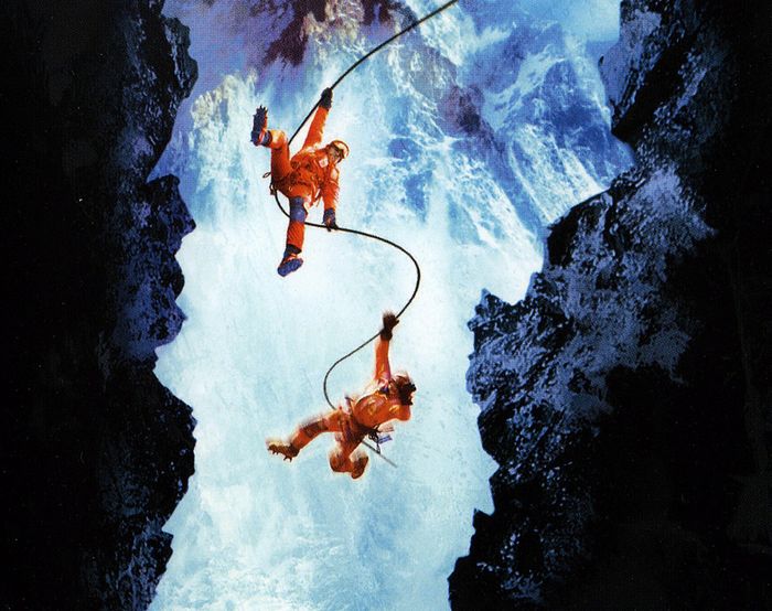 Two climbers wearing bright orange gear and helmets are suspended on a bungee cord between rocky, icy cliffs, above foaming sea waves.