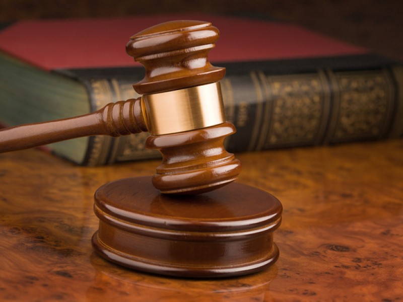 A wooden judge's gavel rests on its sound block on a polished wooden table, with a background of intricately bound law books.