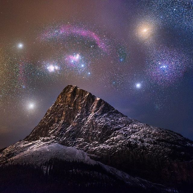 A stunning night sky studded with brightly shining stars and a colorful arc resembling part of the Mazzaroth stretching above a snow-capped, rugged mountain peak.