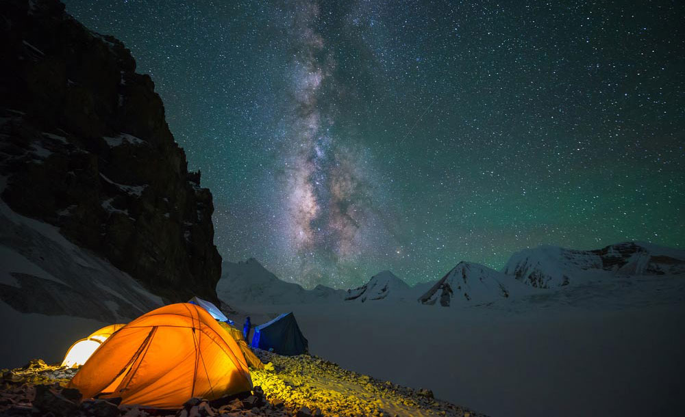 Two illuminated tents nestled on a rugged mountainous terrain under a vast night sky, showcasing the radiant Milky Way and countless stars sprinkled across the heavens.
