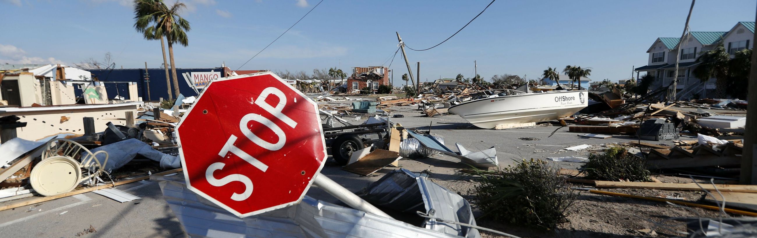 A panoramic view of extensive destruction in a coastal area after a natural disaster. A bent and battered stop sign stands prominently in the foreground amidst scattered debris, including damaged boats, building materials, and a palm tree, suggesting a powerful storm's aftermath.