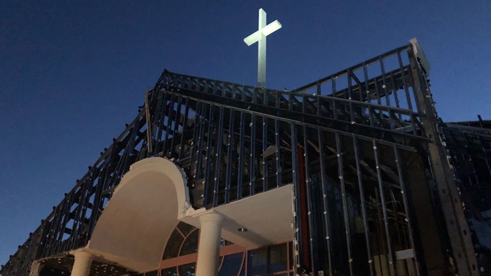 Evening view of an incomplete church building under construction, featuring exposed metal frameworks and arcs. Above the building, a glowing cross is visible against the darkening sky where stars from the Mazzaroth may soon be visible.