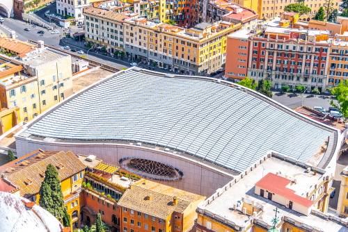 Aerial view of a large, oval-shaped building with a domed, white tiled roof, surrounded by a bustling urban area with multicolored buildings.